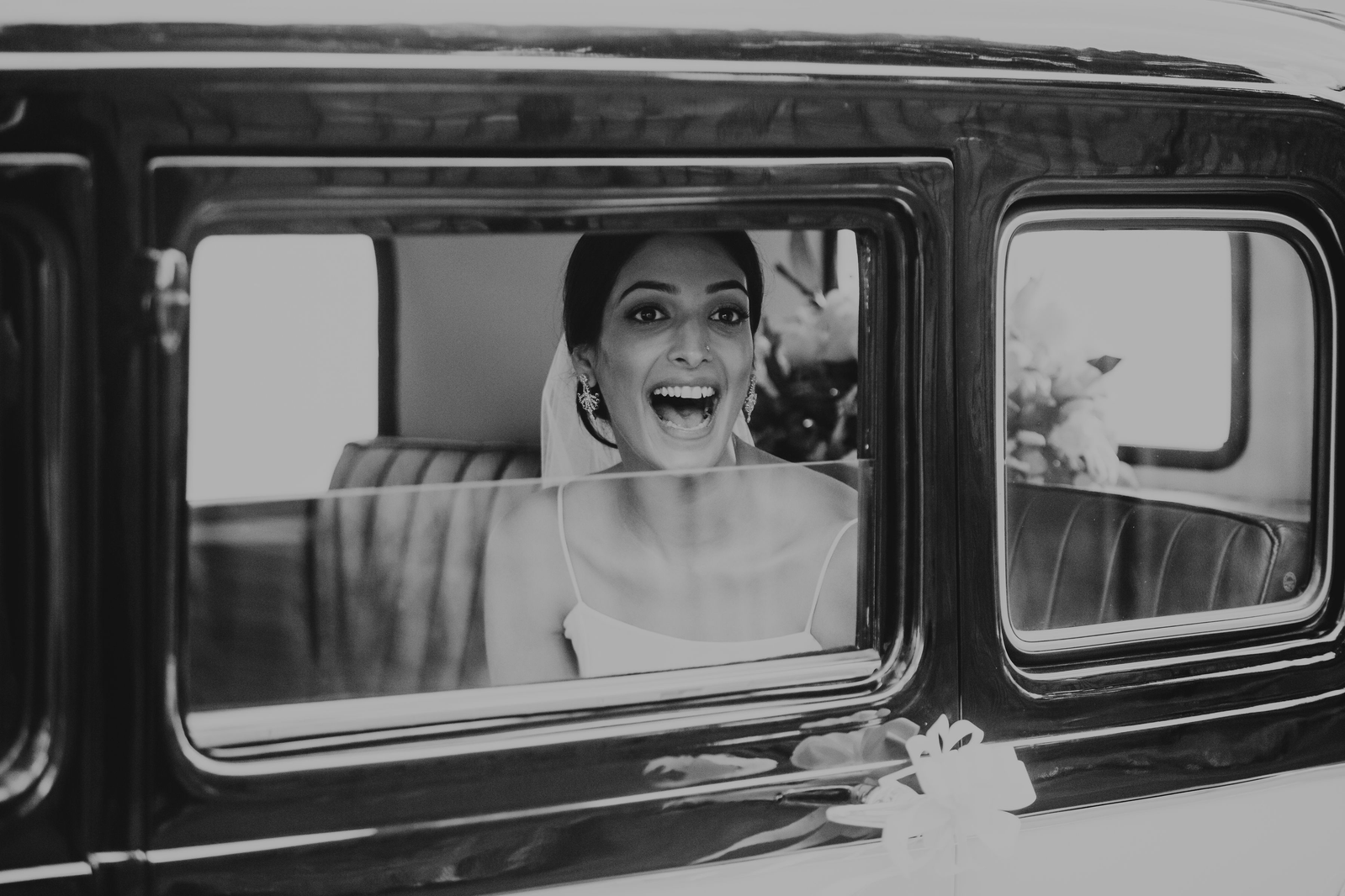 Black and white image of a bride in her wedding car with a huge smile acknowledging wedding guests 