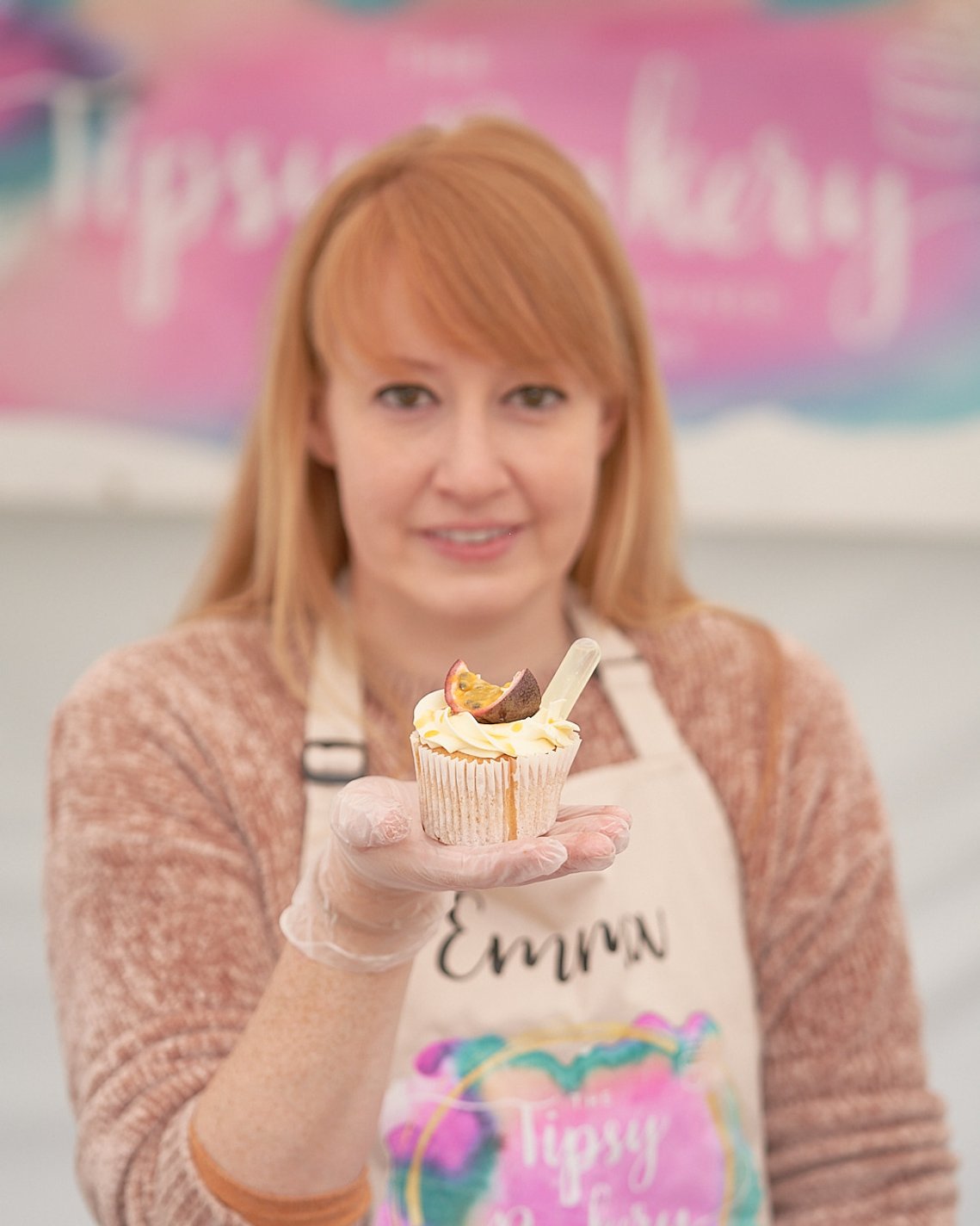 A young woman holding a cupcake out towards the camera. The cake is in focus but the woman is not. Pink background with out-of-focus Tipsy Bakery logo. Brand Photography by Clive Allcorn