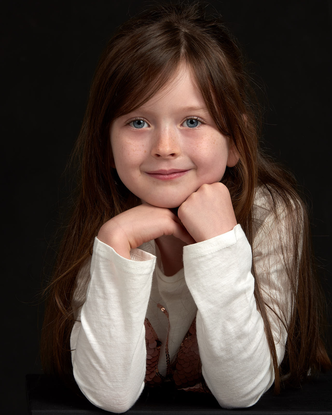 Portrait of a young girl with blue eyes and long brown hair with her chin resting on her hands, looking at the camera. Portrait Photographer - Clive Allcorn