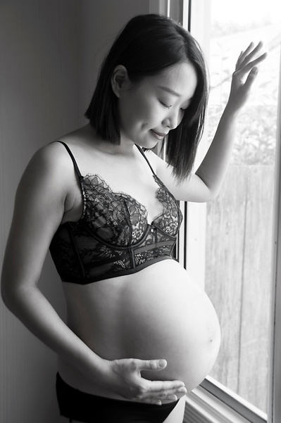 Black and white portrait of a pregnant asian woman wearing lingerie in her bedroom in front of a window