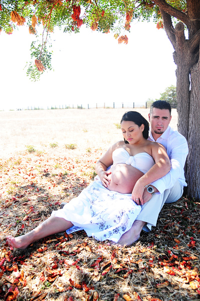 Pregnant woman with her husband wearing white sitting under a tree in the Fall