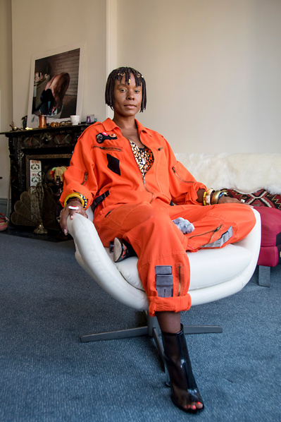 Portrait of black woman in bright orange jumpsuit and short braids sitting on a white chair