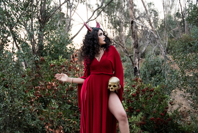 Young woman with dark hair and horns on her head, wearing a high slit red dress, holding a skull
