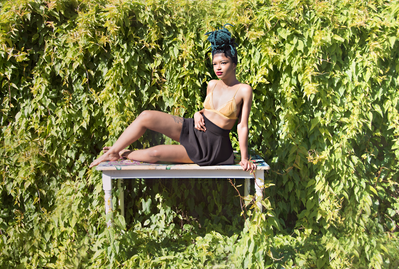 Young black woman with green yarn twists in her hair against a lush overgrown wall of vines on a sunny day