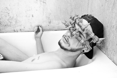 Black and white portrait of a young white man in a bathtub wearing spiked collars over his eyes