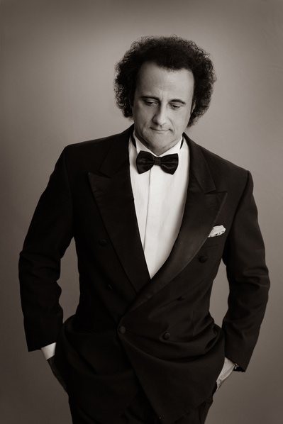 B&W portrait of a male opera singer in a black suit looking down, with hands in his pockets.