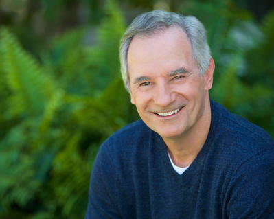 Attractive older male actor outdoor headshot against green leaves wearing Navy sweater.