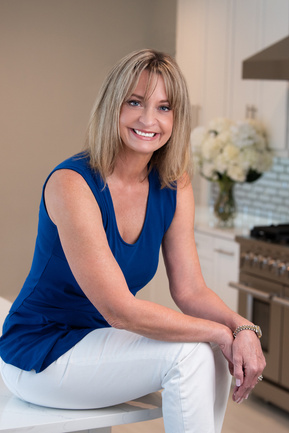 Blond Female kitchen designer seated in beautiful kitchen wearing bright blue top and white jeans