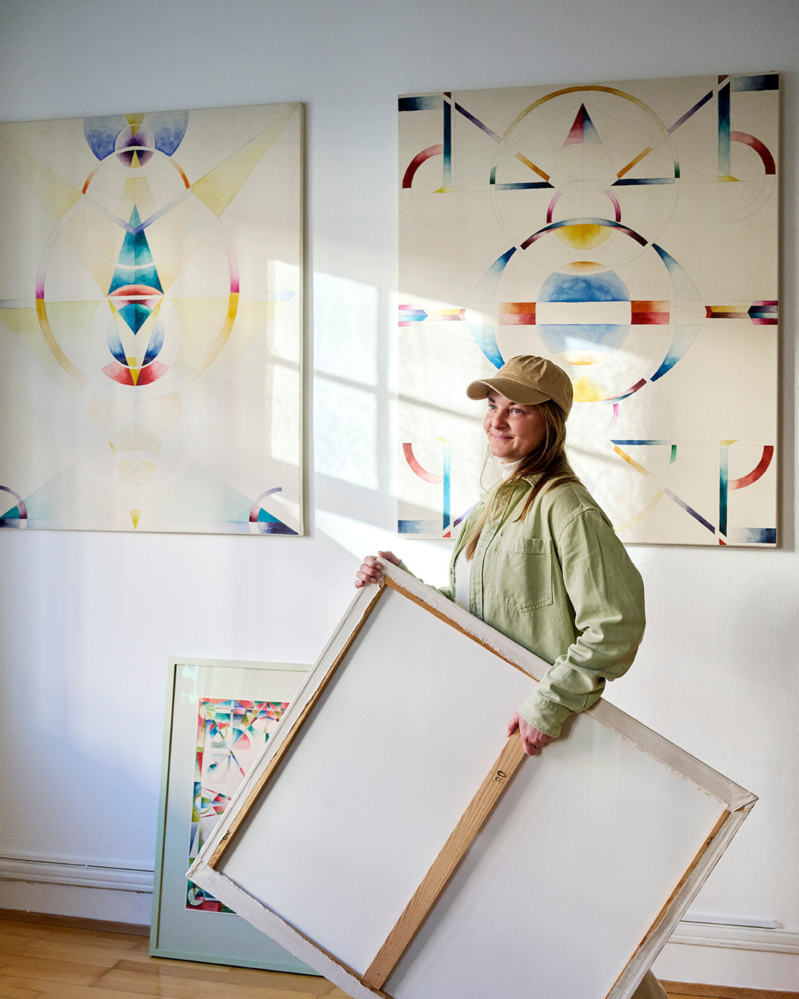 woman moving from right to left, carrying a cavnas, with the back side facing outwards. Two abstract paintings is hanging on the white wall in the background, with a soft light from the windows in the center.