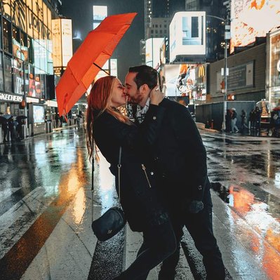 Happy couple holding a red umbrella stand on a cross walk in vibrant American city centre embracing each other tightly and kissing. Show advertising boards lit up behind them.
