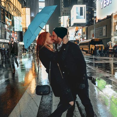 Happy couple holding a blue umbrella stand on a cross walk in vibrant American city centre embracing each other tightly and kissing. Show advertising boards lit up behind them. Image edited to show a rain cloud dropping rain directly on to umbrella.