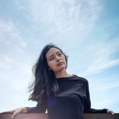 Retouching "after" edit example. A melancholic looking young asian woman looks wistfully away from the camera leant up against a wooden fence in a beautiful rural setting with the sun setting behind her. Photo edited using photoshop retouching.