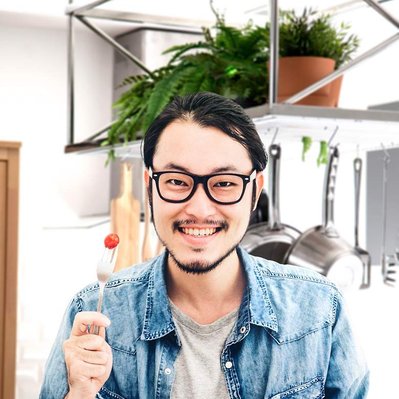 Retouching "before" edit example. A smiling asian male wearing glasses holding a bowl of salad and a fork with a tomato on the end standing in a bright kitchen scene.