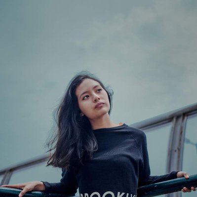 Retouching "before" edit example. A melancholic looking young asian woman looks wistfully away from the camera leant up against a metal fence in a stark urban setting.