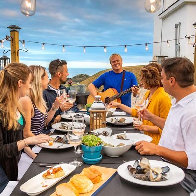 happy guests sit at a table during an outdoor dining experience in Canada watching a young man playing guitar and singing to them. The long table is adorned with various types of delicious looking food including seafood.