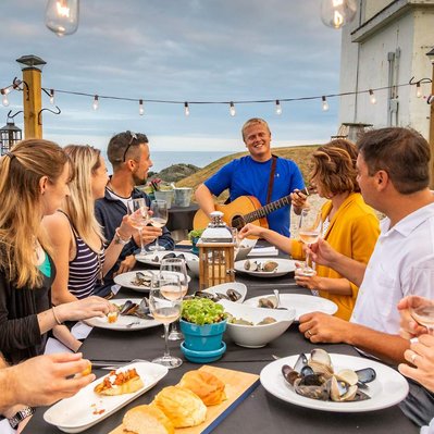 happy guests sit at a table during an outdoor dining experience in Canada watching a young man playing guitar and singing to them. The long table is adorned with various types of delicious looking food including seafood.