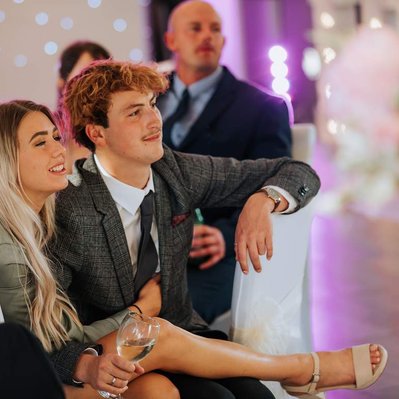 Happy young couple sit close to each other at a wedding reception watching guests dance. Another couple can be seen in the background.