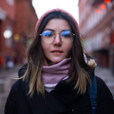 Young Woman wearing glasses with reflections in the lenses stands in a side street alleyway looking directly to camera.