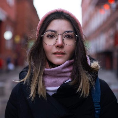 Young Woman wearing glasses with reflections in the lenses removed by Photoshop retouching stands in a side street alleyway looking directly to camera.
