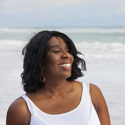 Retouching "after" edit example. A black  female on a beach smiling, with the ocean behind her. The man has been removed using Photoshop retouching.