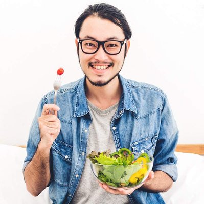 Retouching "before" edit example. A smiling asian male wearing glasses holding a bowl of salad and a fork with a tomato on the end.