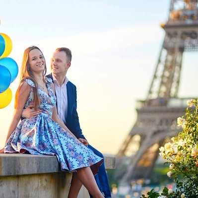 Retouching "before" edit example. A happy caucasian couple holding a bunch of balloons smile and pose for a photo sat on a wall with the eiffel tower in the background with sun setting.