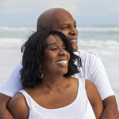 Retouching "before" edit example. A black male and female couple stand closely on a beach smiling, with the ocean behind them.