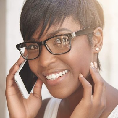 Beautiful young black woman with short dark hair smiling, wearing glasses (with reflections) whilst holding mobile phone to her ear and looking directly at the camera, backlit with warm sunlight.