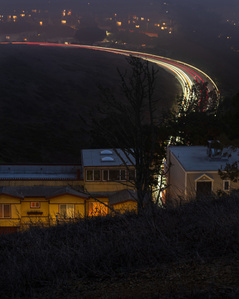 A long exposure shot of cars driving up and down Twin Peaks, leaving light trails in their wake as they drive by two homes that can be seen glowing through the fog.