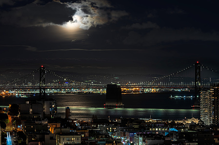 The full moon, obscured by clouds, rises over the San Francisco-Oakland Bay Bridge, with Oakland in the background and San Francisco in the foreground.