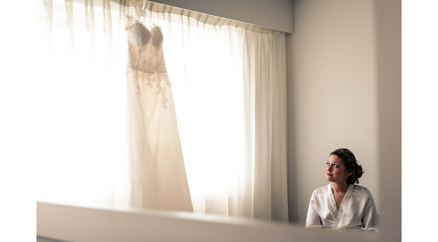 Chica con bata de satén mira su vestido de novia colgado en la ventana