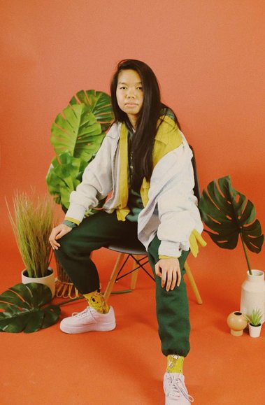 Allison Masangkay (aka DJ Phenohype) sitting in a chair wearing an outfit of yellows, greens, lavender, and light wash denim blue with a warm orange backdrop, surrounded by several green plants. Photo by Bianca Recuenco (@biancarecuenco).