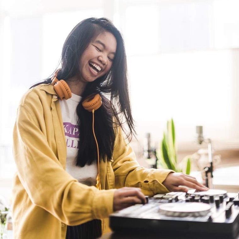 Allison Masangkay, a Filipinx femme with light tan skin and long black hair, smiles with their eyes slightly closed looking down at their DJ controller. They wear orange headphones, a white and lavender t-shirt, and a yellow long-sleeve button-up shirt.