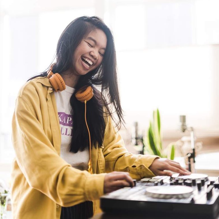 Allison Masangkay, a Filipinx femme with light brown skin and long black hair, smiles with their eyes slightly closed looking down at their DJ controller. They wear orange headphones, a white and lavender t-shirt, and a yellow long-sleeve button-up shirt.