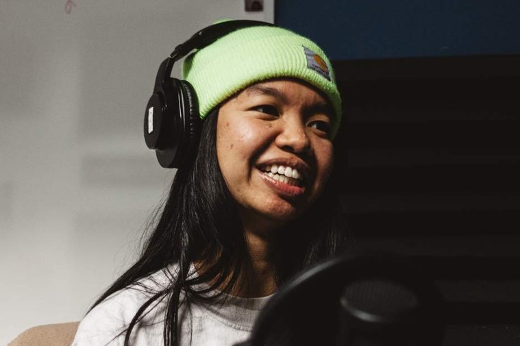 Allison Masangkay, a Filipinx femme with long black hair, wearing a white shirt and yellow beanie with black headphones and studio microphone