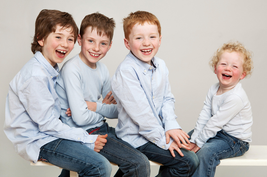 Professional family photo of four brothers, young boys in blue shirts smiling and laughing sitting on bench in photography studio Dublin