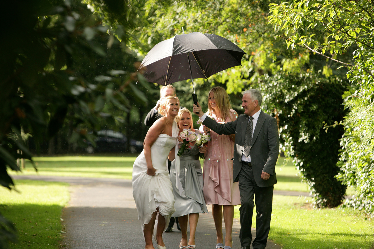 Wedding day photos Bride and bridesmaids with Father of the bride huddled under an umbrella in the park fun reportage documentary style wedding photography