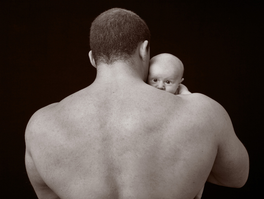 A black and white portrait of a male body builder holding a tiny newborn baby on his shoulder. Taken in a professional family portrait photography studio in Dublin with a black background 