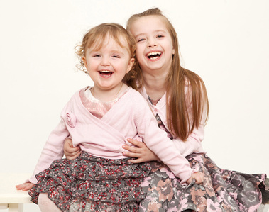Two sisters laughing and hugging sitting on a bench in professional family photography portrait studio