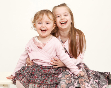 Two sisters laughing and hugging sitting on a bench in professional family photography portrait studio