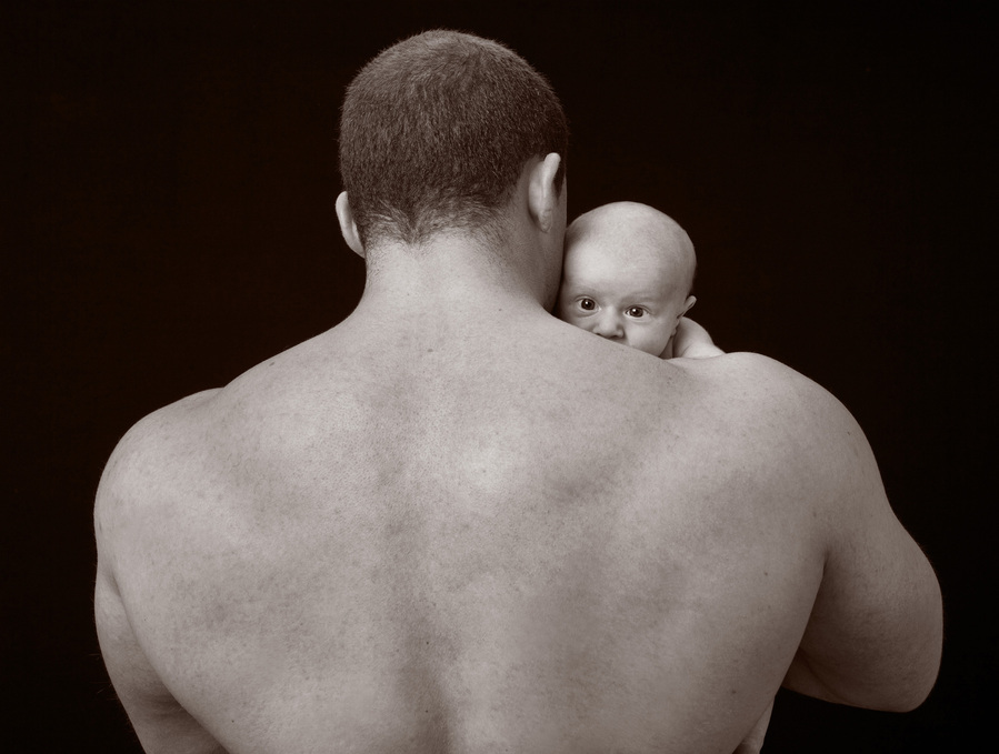Black and white professional studio portrait of a male bodybuilder with a tiny newborn over his shoulder taken in a family portrait photography studio in Dublin 