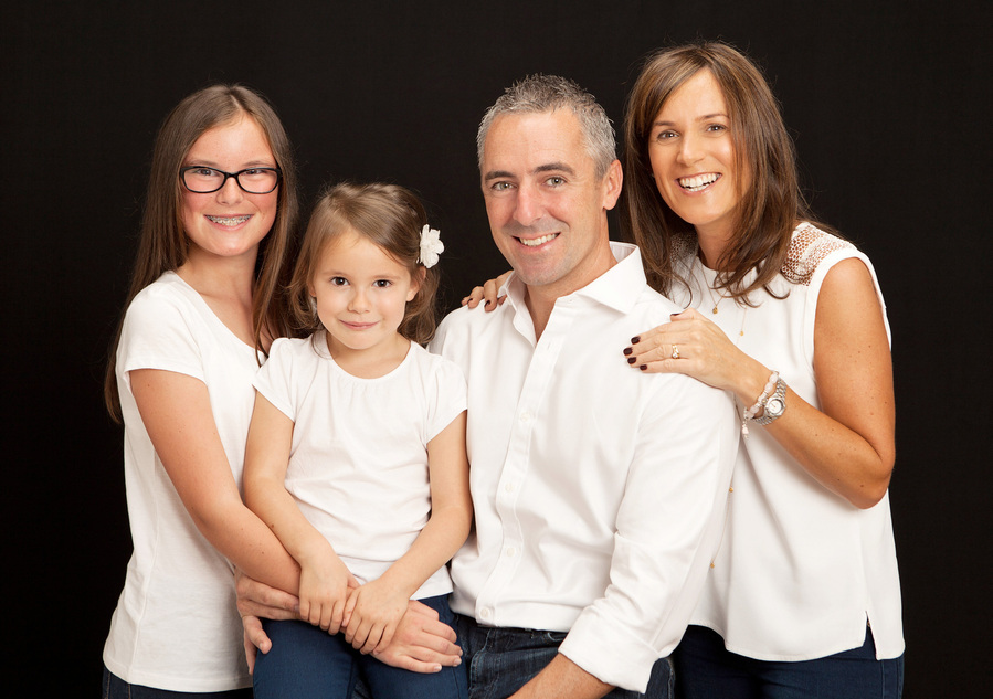 Family portrait of Mother, Father and two daughters wearing white shirts. Taken in a professional photography studio with a black background