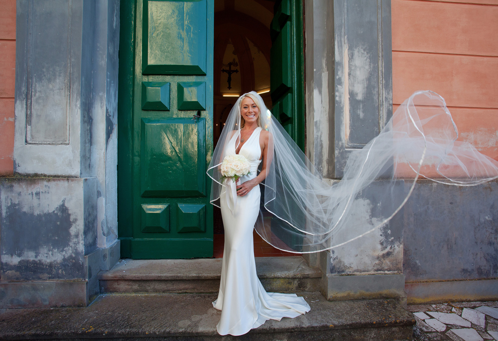 Smiling blonde bride with veil blowing in front of large green doors Location wedding Italy relaxed fun style wedding photography