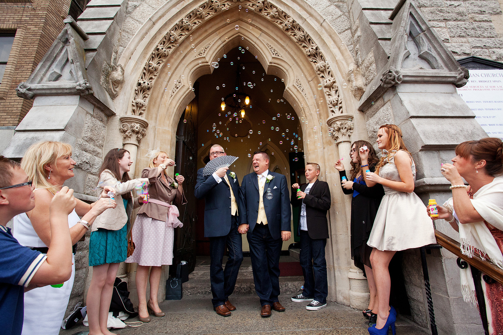 Newly married groom and groom leaving church congregation blowing bubbles on gay couple reportage documentary style wedding photography
