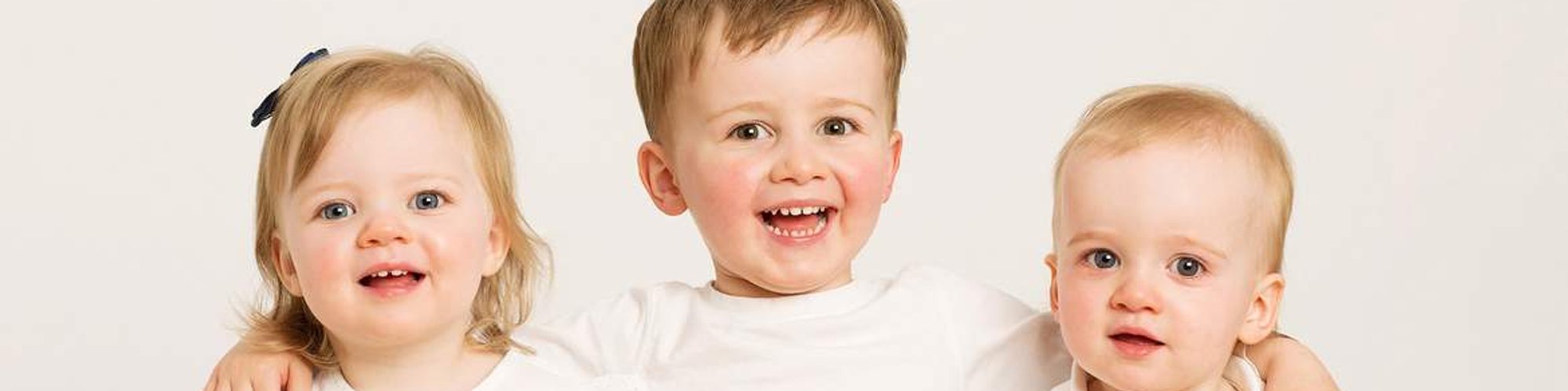 Classic family portrait of 3 young children with arms around each other wearing white and denim in professional photography studio Dublin 