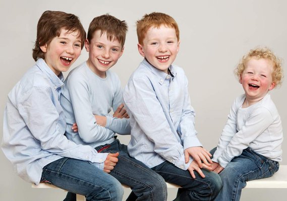 Professional family photo of four brothers, young boys in blue shirts smiling and laughing sitting on bench in photography studio Dublin 