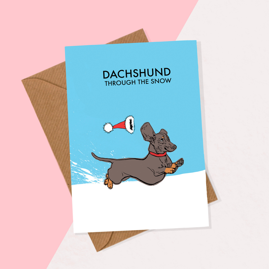 Dachshund through the snow.  Christmas card by Woof Portraits and Ryan Hodge illustration.  Blue background, Santa hat, running dog. Recycled paper envelope. 
