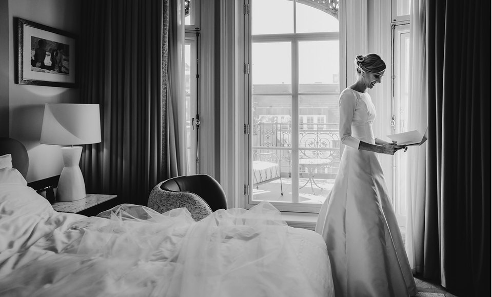 A bride in her wedding dress  standing by the window of her hotel room. She is reading piece of paper in her hand