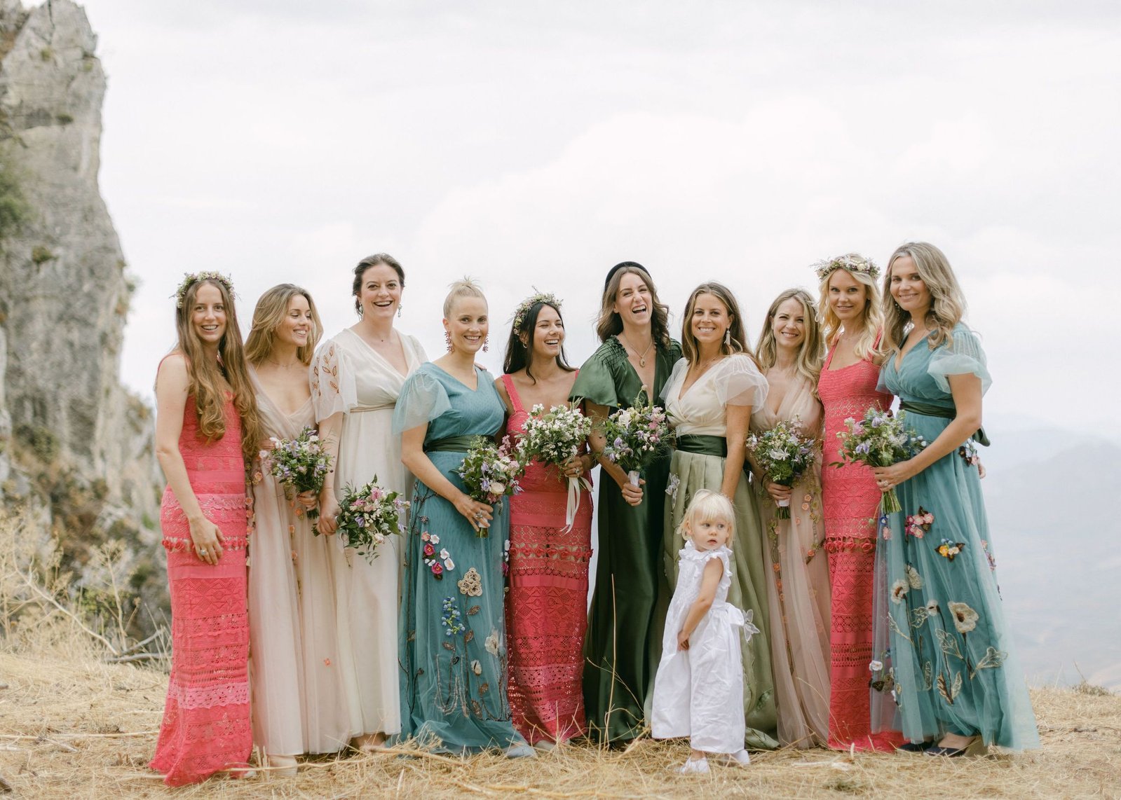 Ten bridesmaids stand together with a blonde haired flower girl on a hilltop in Sicily. They are all holding a bouquet of flowers each smiling into the camera.