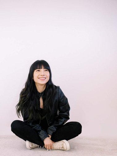 Asian woman with soft wavy hair, wearing leather jacker,  sitting on the ground smiling.